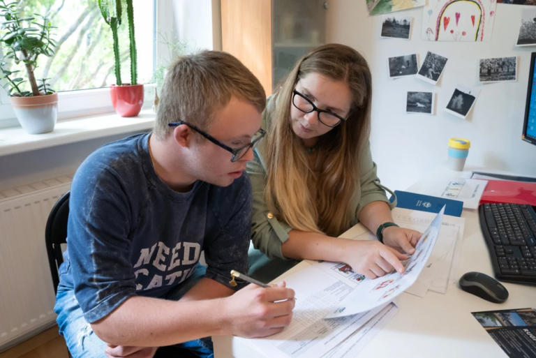 Vaikas sėdi su mokytoja ir kąžką rašo.