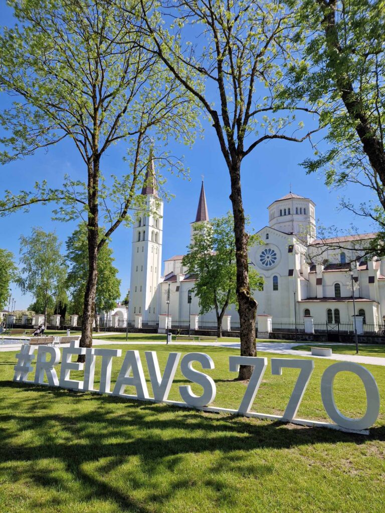 Nuotraukoje yra balta bažnyčia, o nuotraukos priekyje, ant žolės, yra padėtos raidės #RĖITAVS770.