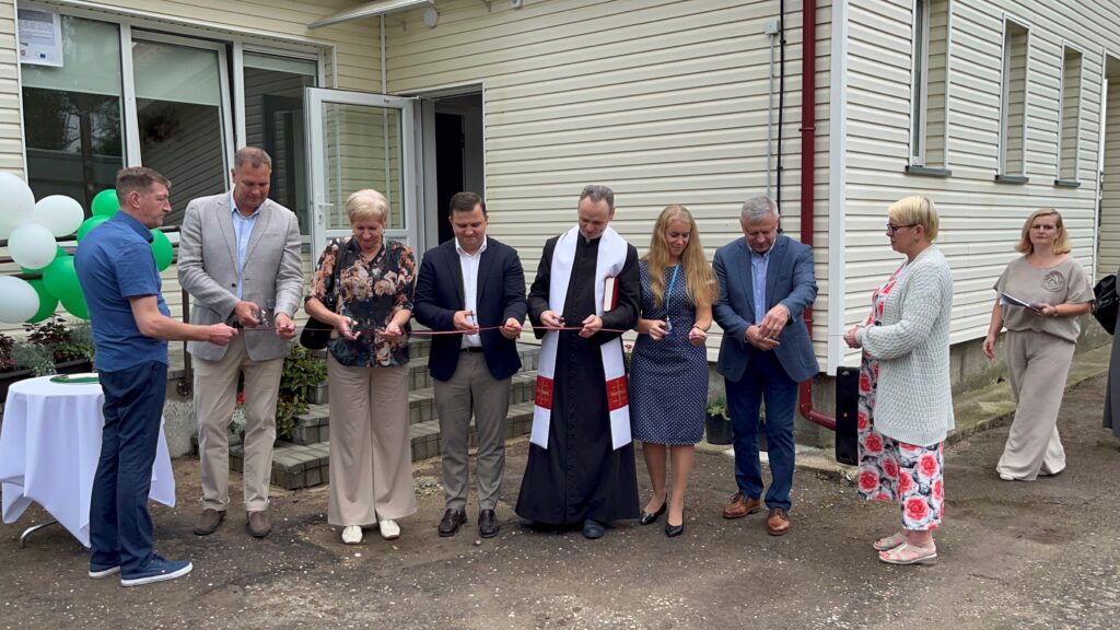 Nuotraukoje yra 9 žmonės prie namo lauke. Visi laiko juostą ir ją kerpa, taip simboliškai atidarydami grupinio gyvenimo namus. Nuotraukoje yra tinklaveikos specialistė Marijona Janavičienė, Šilalės rajone dirbantis kunigas ir kiti žmonės. 