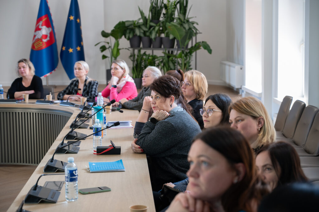 Nuotraukoje prie stalo sėdi susitikimo dalyviai iš įvairių įstaigų ir klausosi pranešimų.