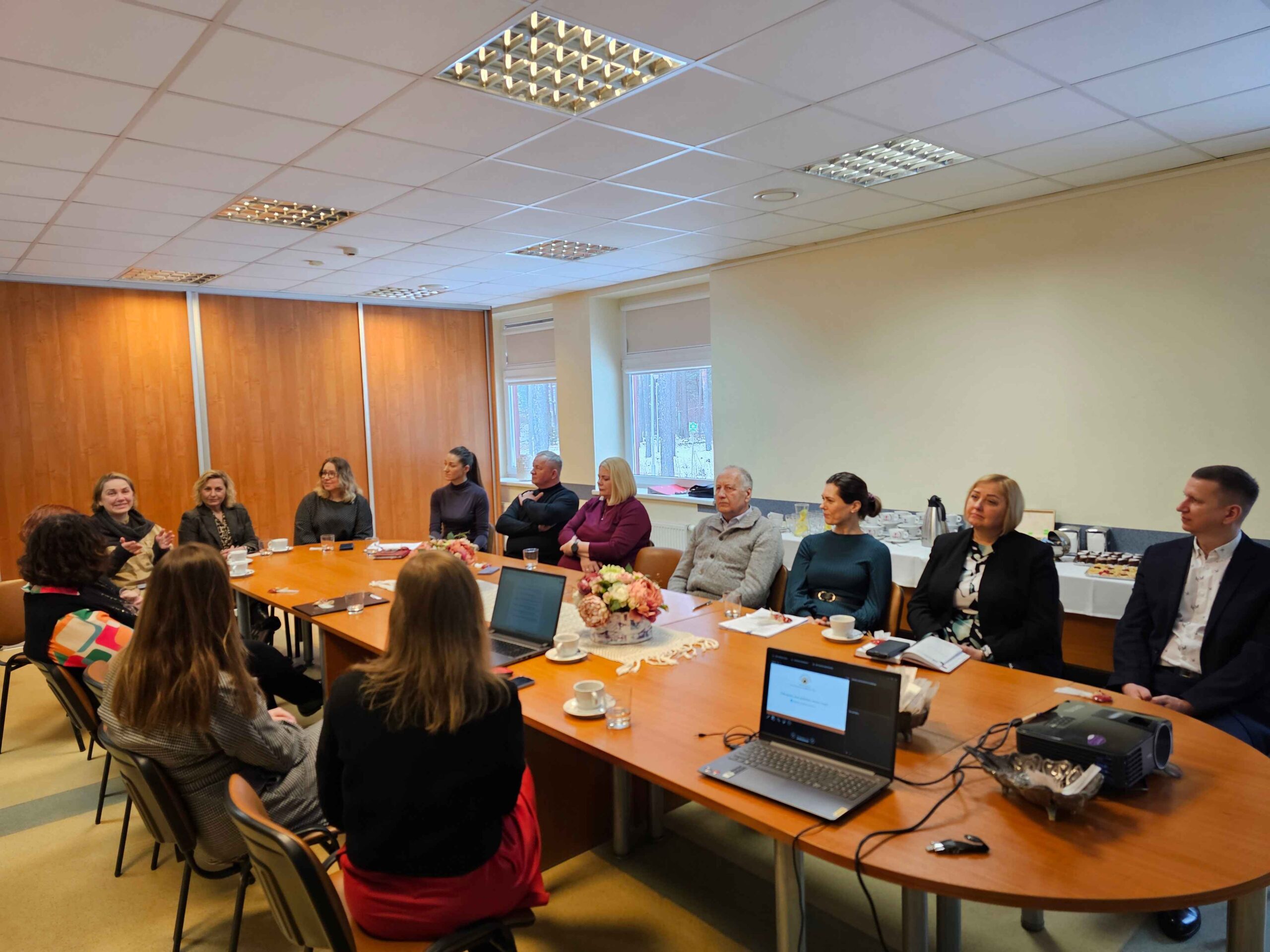 Salėje prie didelio stalo sėdi apie 20 žmonių, dauguma jų moterys. Ant stalo yra nešiojami kompiuteriai, užrašų knygos, stalo priekyje padėtas vaizdo projektorius.