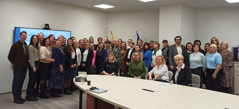 Nuotraukoje matoma didelė grupė žmonių, daugiausia moterys, bet yra ir keli vyrai. Jie pozuoja bendrai nuotraukai modernioje konferencijų salėje. Dalis žmonių stovi, o keletas moterų sėdi prie šviesaus ovalo formos stalo. Visi žiūri į kamerą, kai kurie šypsosi, kiti turi rimtus ar neutraliai draugiškus veido išraiškas. Už grupės yra didelis ekranas, kuriame matoma dalis užrašo „Keisti ir keistis“, o šalia – dvi vėliavos, viena iš jų – Europos Sąjungos. Aplinka šviesi ir moderni, su minimalistiniu dizainu, būdinga oficialiems renginiams ar seminarams. Ant stalo matyti nešiojamas kompiuteris, stiklinė vandens ir keletas dokumentų. Bendra nuotraukos atmosfera yra profesionali, bet šilta – žmonės atrodo bendruomeniški, dalyvaujantys dalykiniame, bet draugiškame renginyje.