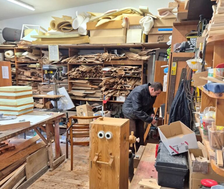 Kambaryje yra daug medinių spintų, visos jos pilnos medinių lentų ar medinių dalių. Ant spintų viršaus taip pat pridėta daiktų, daugiausia medžiagų. Prie vienos spintos stovi šiek tiek pasilenkęs žmogus, kažko ieško spintoje.