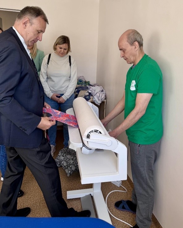 Prie rūbų lyginimo staklių stovi vyras juodu kostiumu, už staklių stovi vyras, kuris yra socialinių dirbtuvių lankytojas, jis dėvi žalius marškinėlius, skalbyklos uniformą. Vyrus skiriančios staklės yra sudarytos iš pailgo ritinio, kuris sukdamasis slegia audinį ir taip jį išlygina. Šioje akimirkoje naudojamas margas audinys. Iš šalies visą lyginimo procesą stebi susidomėjusi moteris.