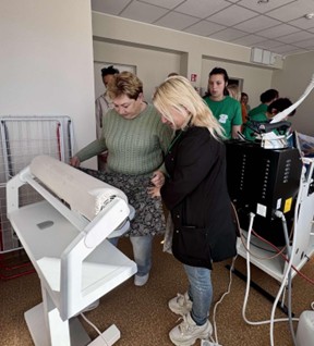 Dvi moterys stovi prie lyginimo staklių. Kairėje stovinti moterys laiko margą audinį, rodo kitai moteriai lyginimo eigą.