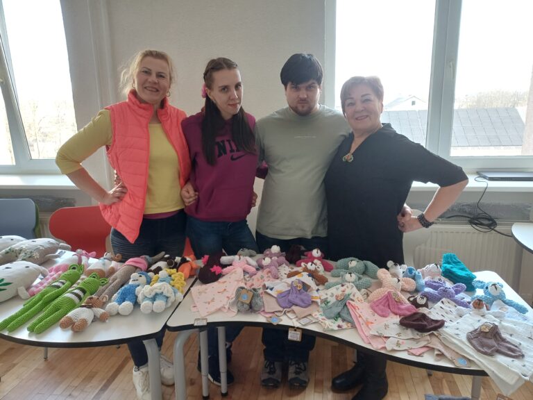 Ant stalo sudėti minkšti žaislai ir maži audiniai. Už stalo stovi 4 žmonės (3 moterys ir vyras), jie susikabinę ir žiūri į fotografuojančią kamerą, šypsosi.