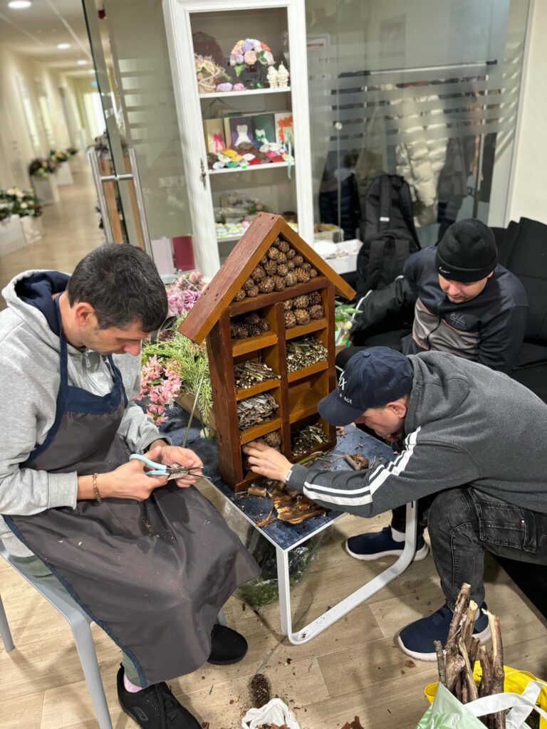 Kambaryje ant žemo stalo yra namo formos medinė dežutė su stalčiais. Aplink yra trys vyrai, vienas iš jų deda į dėžutę konkorežį, kitas sėdi ant sofos ir žiūri, kitas kerpa siūlą su žirklėmis.