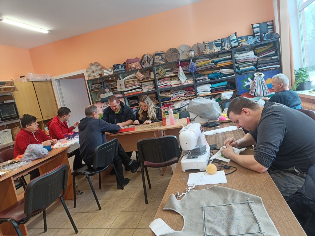 Kambaryje yra daug stalų, prie jų sėdi žmonės. Ant stalų yra siuvimo mašina, audiniai, įvairūs įrankiai. Prie stalo sėdintys žmonės dirba su rankomis. Prie kambario sienos yra didelė spinta, pilna audeklų.