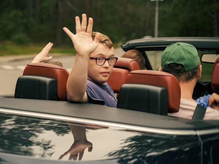 Vaikinas sėdi automobilio be stogo galinėje sėdynėje, persisuskęs į galą moja ranka.