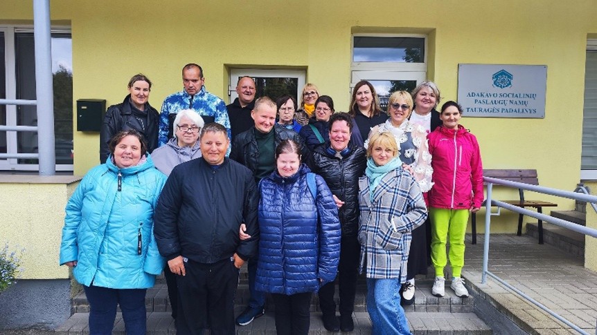Lauke stovi socialinių dirbtuvių lankytojai, jie šypsosi, džiugiai nusiteikę. Jie yra prie pastato, ant kurio yra lentelė: Adakavo socialinių paslaugų namų Tauragės padalinys.