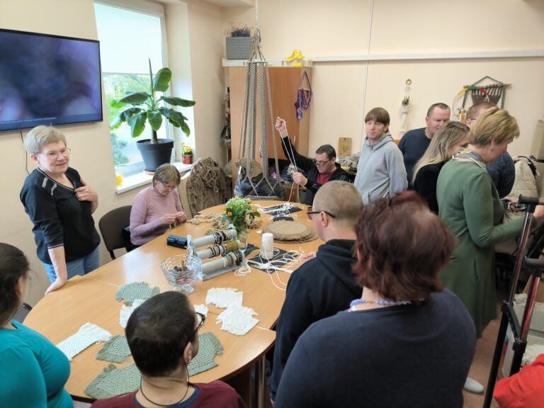 Prie stalo sėdi daug žmonių, ant stalo daug nėrinių, yra sudėtos apyrankės. Šalia stalo nuo sienos kabo dar vis daroma besisupanti kėdė. Šalia yra stovintys žmonės, jie kalbasi.