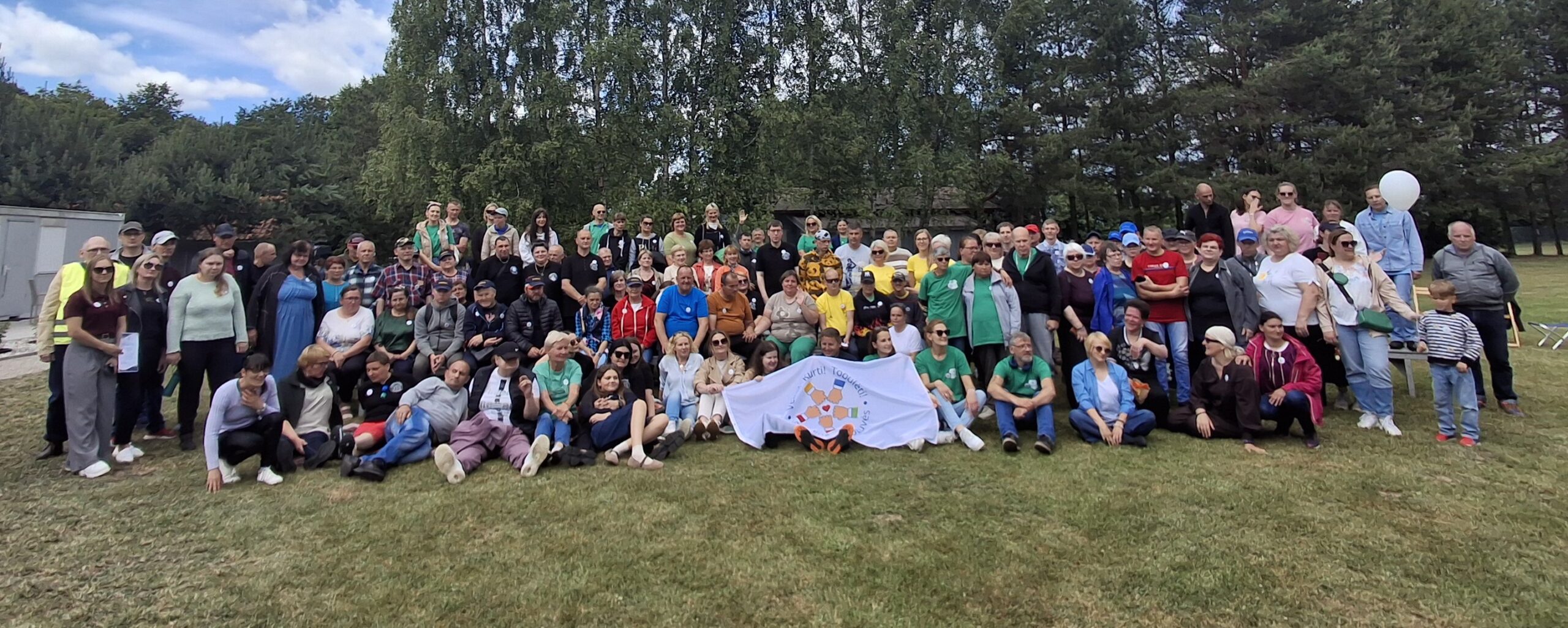 Lauke ant vejos keliomis eilėmis sustoję ir susėdę socialinių dirbtuvių dalyviai ir svečiai. Glaudžiai stovi, dalis apsikabinę. Pirmoje eilėje žmonės laiko vėliavą su logotipu, kuriame vaizduojamos penkios rankos suspaustos į kumščius ir ratu jungiančios viena kitą. Aplink ženklą ratu eina šūkis: Veikti, kurti, tobulėti.