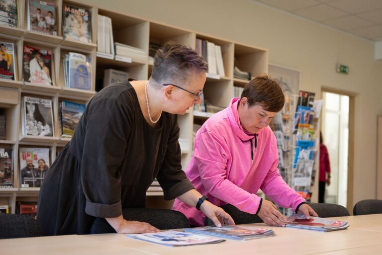 Bibliotekininkė DUOday dalyvei Ingridai rodo ant stalo esančius žurnalus. Moterys yra bibliotekoje, stovi prie stalo ir peržiūri žurnalus, už jų prie sienos esanti komoda yra pilna žurnalų ir knygų.