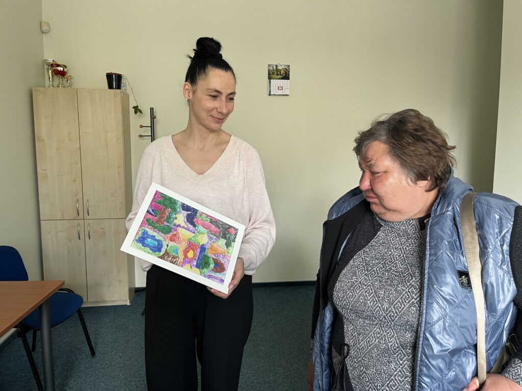 Pagalbos priimant specialistė Viktorija rodo DUOday dalyvei Vilmai piešinį, gautą iš kliento. Moterys yra kambaryje, kurio šone yra komoda, matosi stalas ir kėdė.