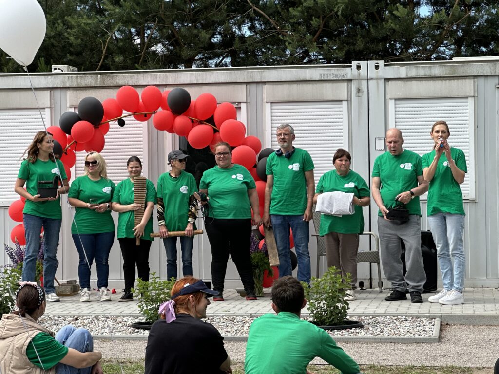 Iš arti ant scenos su balionais stovi žaliais marškinėliais pasipuošę Jurbarko socialinių dirbtuvių skalbykla kolektyvas. Visi jie laiko po vieną skalbimui reikalingą įrenginį - kas medines kultuves, lininius audinius ar lygintuvus.