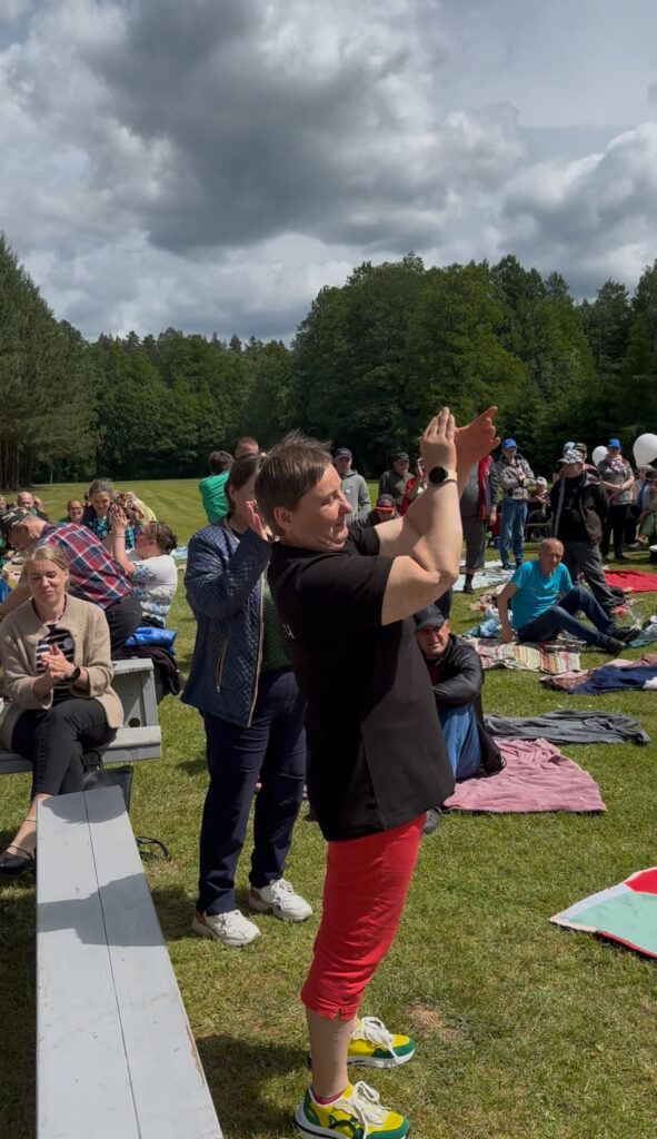 Pievoje atsistojusi ploja laiminga moteris, tolumoje ploja ir kiti socialinių dirbtuvių nariai.