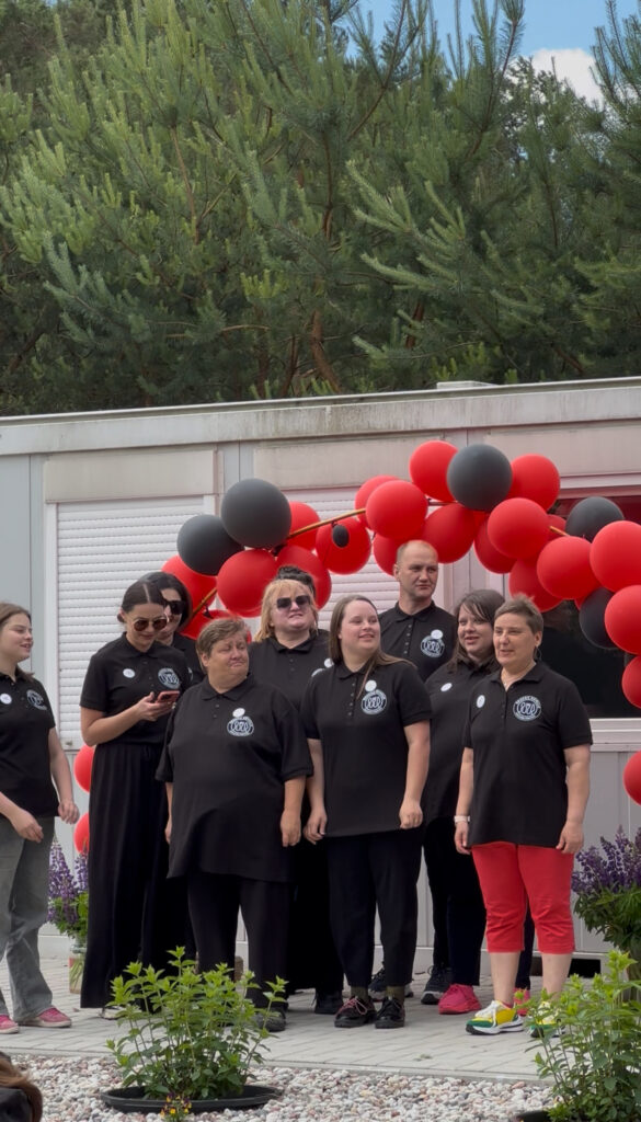 Iš arti, iš Tauragės atvykusio socialinių dirbtuvių Draugų kiemelis kolektyvas. Juodais maškinėliais pasipuošę komandos nariai yra atsisukę į dešinėje kalbančią atstovę. Už jų yra balionų arka.