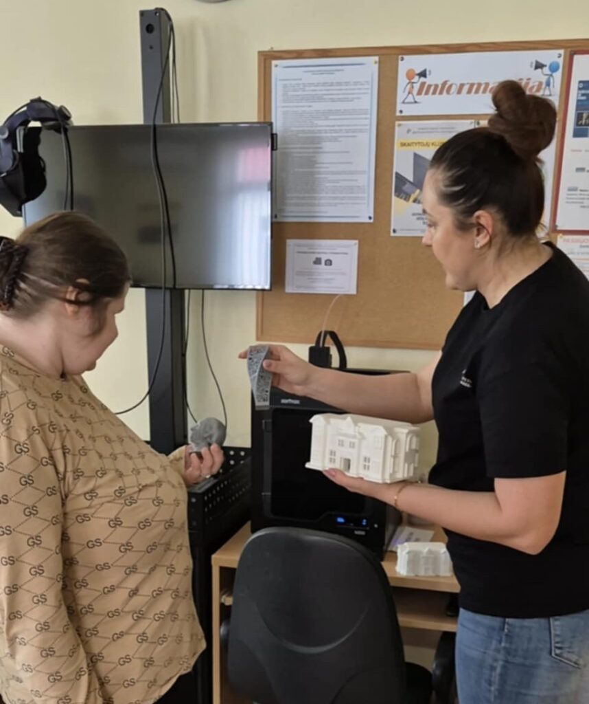 Bibliotekos darbuotoja rodo DUOday dalyvei Jurgitai 3D spausdintuvą. Ant stalo yra daug technikos, didelis ekranas, o ant sienos yra skelbimų lenta su keliais skelbimais.