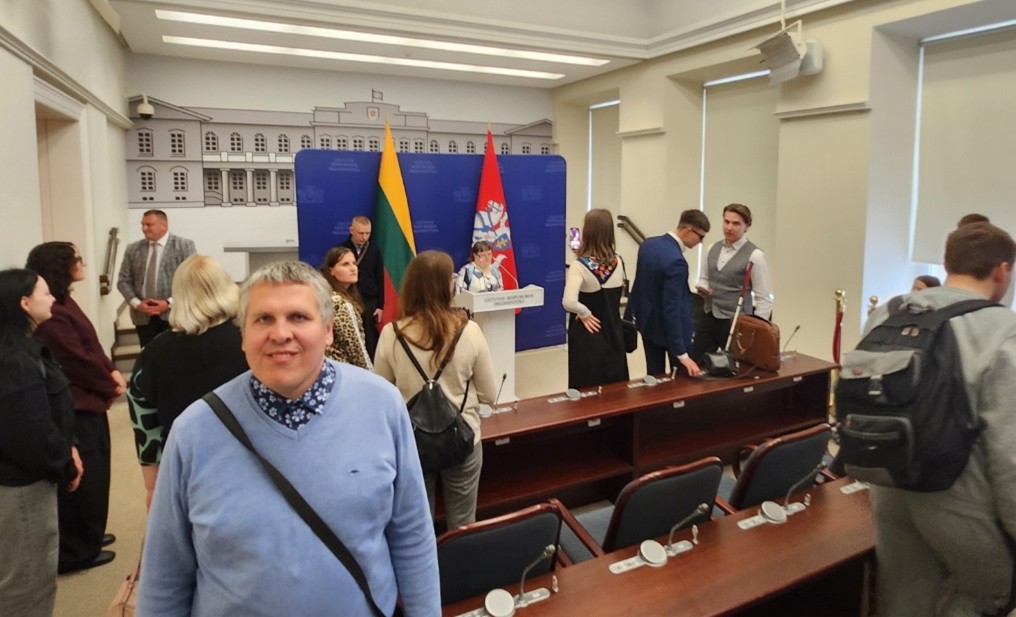 Socialinių dirbtuvių dalyvis Adolfas stovi pranešimų spaudos salėje Prezidentūroje. Salės priekyje yra sienelė, kalbėjimo pakyla su mikrofonu, šalia pastatyta Lietuvos vėliava ir herbas. Salėje yra stalai su kėdėmis. Salėje vaikšto ir fotografuojasi žmonės, kiti DUOday iniciatyvos dalyviai.