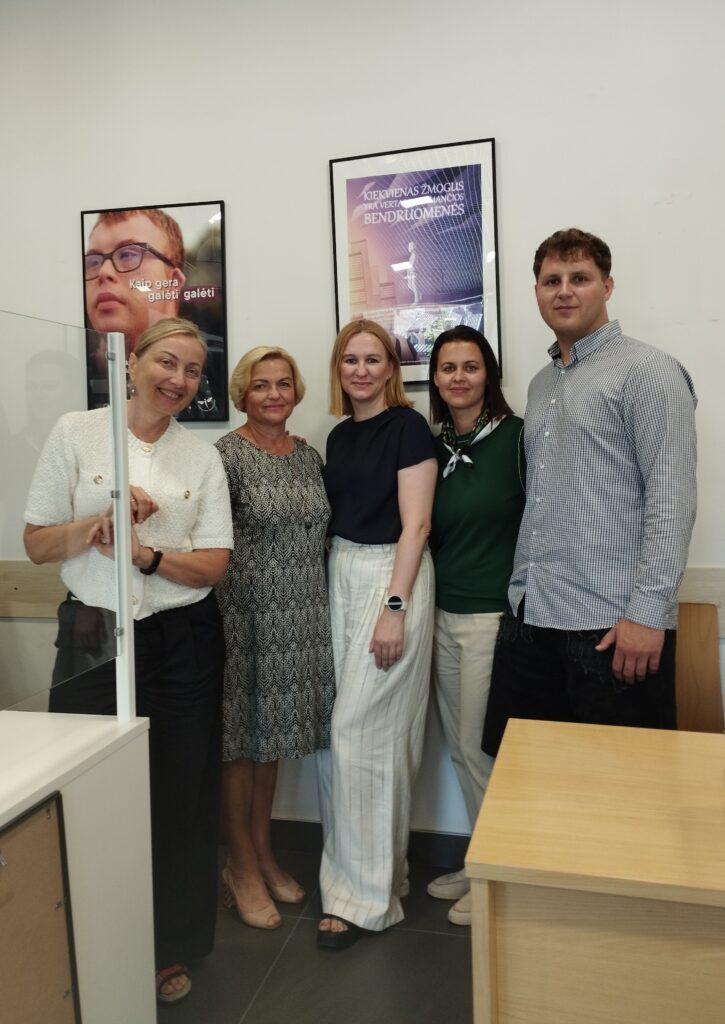 Nuotraukoje užfiksuota kaip susitikimo dalyviai (iš kairės į dešinę) Asmens su negalia teisių apsaugos agentūros direktorė Eglė Čaplikinė ir Kanclerė Vytautė Polujanskienė, Pertvarkos projekto vadovė Svajonė Rainienė ir tinklaveikos specialistė Vilniaus regionui Kristina Maciūtė-Stukanė bei Elektrėnų savivaldybės Socialinės paramos skyriaus atvejo vadybininkas Edvinas Gurskas stovi ir šypsosi pozuodami. Už jų matomi du įrėminti plakatai su užrašais: „Kaip gera galėti galėti“ ir „Kiekvienas žmogus yra vertas jį priimančios bendruomenės“. 
