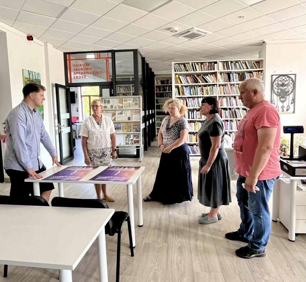 Nuotraukoje penki suaugę žmonės stovi bibliotekoje arba informacijos centre šviesioje erdvėje. Vienas vyras stovi prie balto stalo ir rodo du plakatus, padėtus ant stalo. Už nugaros – lentynos pilnos knygų, ant stiklinių durų užrašas „Vaikų ir jaunimo aptarnavimo erdvė“. Kiti keturi asmenys klauso ir žiūri į plakatus. Aplinka rami ir intelektuali.