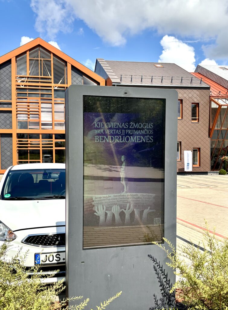 Nuotraukoje lauke prie modernaus pastato su oranžiniais architektūriniais elementais stovi elektroninis informacinis stendas. Jo ekrane rodomas plakatas su žinomu šūkiu: „Kiekvienas žmogus yra vertas jį priimančios bendruomenės“. Fone matoma dalis pastato, balta mašina, žolė ir dangus su debesimis. 