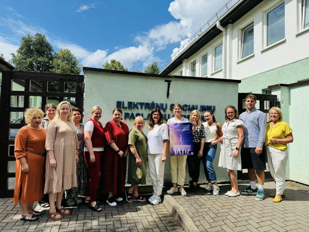Nuotraukoje didelė grupė žmonių pozuoja lauke prie pastato, ant kurio fasado parašyta „Elektrėnų socialinių paslaugų centras“. Grupė laiko minėtą plakatą apie bendruomenę. Visi šypsosi, atmosfera draugiška. Dangus giedras, vasariška diena.