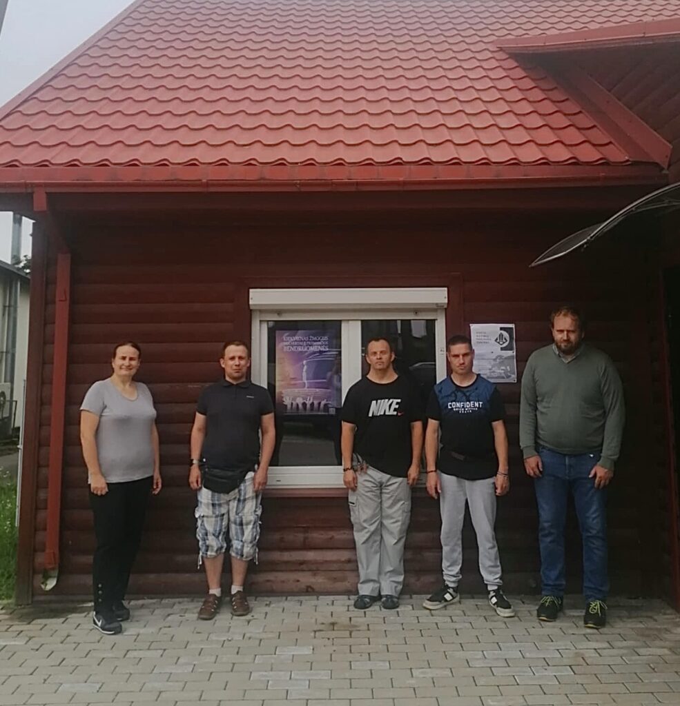 Penki socialinių dirbtuvių dalyviai stovi lauke prie medinio pastato su raudonu stogu. Ant lango užklijuotas tas pats violetinis plakatas. Grupė išsirikiavusi tvarkingai, žiūri į kamerą. Apranga laisvalaikio, fonas – trinkelėmis grįstas kiemas.
