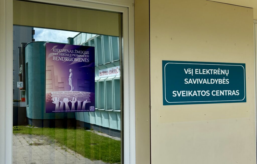 Nuotraukoje plakatas su užrašu „Kiekvienas žmogus yra vertas jį priimančios bendruomenės“ priklijuotas ant lango, šalia įėjimo į pastatą su iškaba „VšĮ Elektrėnų savivaldybės sveikatos centras“. Langas atspindi priešais esantį žalią pastatą. Vaizdas dvelkia oficialumu, aplinka tvarkinga.