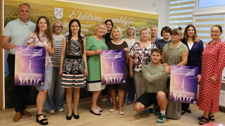 Nuotraukoje didelė grupė žmonių, tarp jų vyrų ir moterų, pozuoja Elektrėnų savivaldybėje. Jie laiko tris plakatus su tuo pačiu bendruomeniškumą skatinančiu šūkiu. Už nugaros geltona sienelė su Elektrėnų savivaldybės logotipu. Visi atrodo įsitraukę, atmosfera profesionali, bet draugiška.