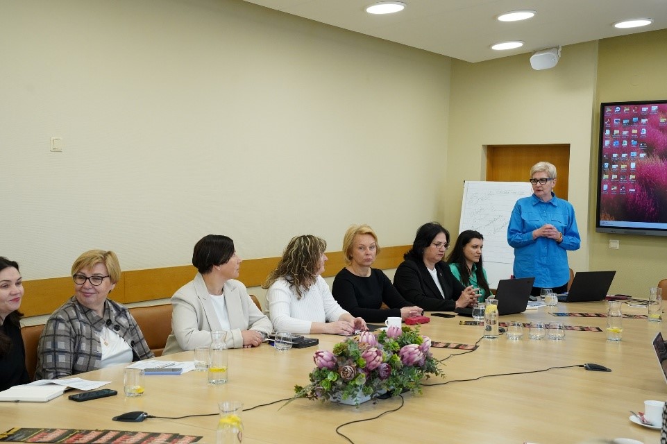 Salėje, prie didelio stalo sėdi 7 moterys, o stalo priekyje pranešimą skaito Marijona Janavičienė. Šalia jos yra užrašų lenta ir interaktyvi lenta, ant kurios matosi sujungto kompiuterio ekranas.