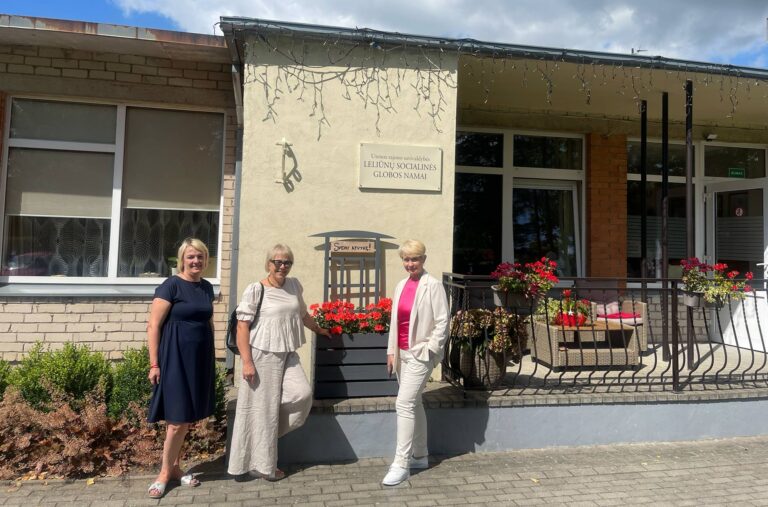 Prie pastato stovi trys besišypsančios moterys. Ant pastato kabo lentelė su užrašu "Utenos rajono savivaldybės Leliunų socialinės globos namai"