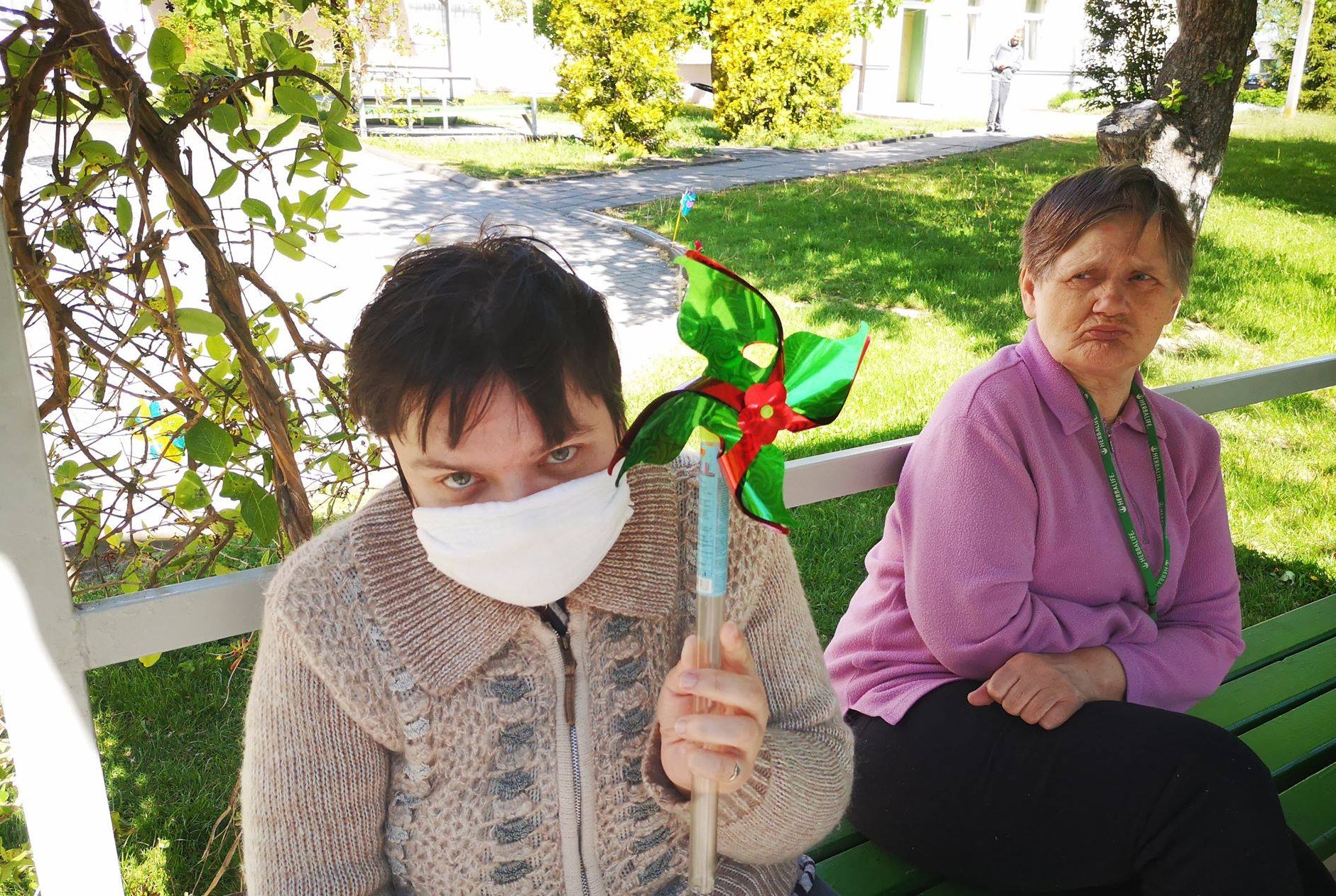 Dvi moterys sėdi lauke, viena iš jų su medicinine kauke, rankoje laiko vėjo malūnėlį. Lauke šilta, aplink žaliuoja žolė.