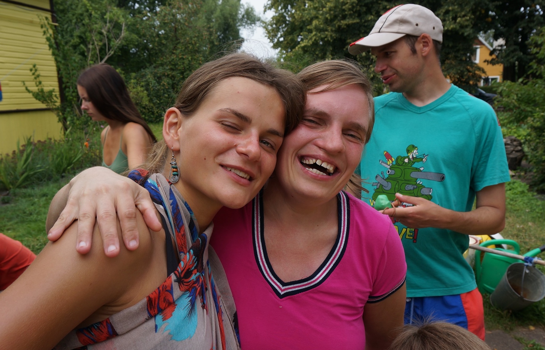 Dvi moterys apsikabinusios, šypsosi. Jos yra lauke, šalia yra medžiai, kiti žmonės, vasara.