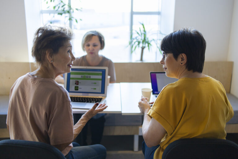 Dvi moterys sėdi prie stalo, kiekviena jų turi nešiojamą kompiuterį, kurie yra padėti ant stalo prieš jas. Moterys tarpusavyje kalbasi.