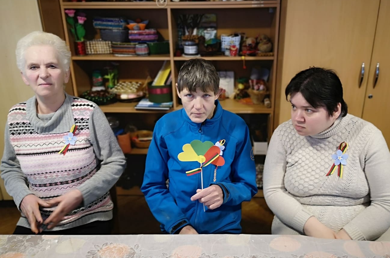 Kambaryje prie stalo yra trys moterys su intelekto ar psichosocialine negalia, rankose turi Lietuvos atributiką.