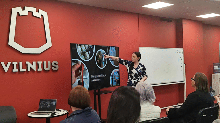 Savivaldybės salėje moteris skaito pranešimą. Ji rodo į ekraną, kuriame yra parašyta „nauji produktai ir paslaugos“, ekrane taip pat yra kelios žvakių, socialinių dirbuvių gaminamų daiktų nuotraukos. Priešais ekraną sėdi žmonės ir žiūri į pranešimą skaitančią moterį.