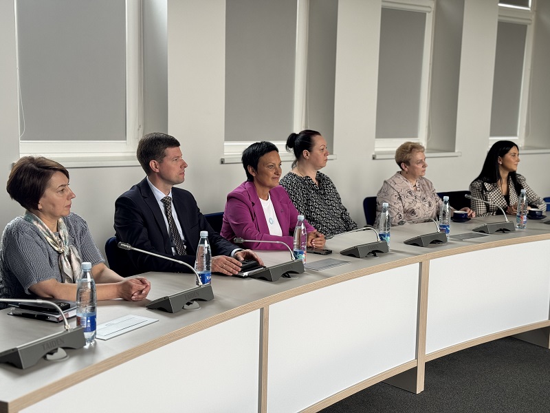 Nuotraukoje šeši žmonės sėdi prie išlenkto konferencijų stalo, prieš kiekvieną padėtas mikrofonas ir vandens butelis. Aplinka oficiali, su dideliais langais ir dalinai užtrauktomis žaliuzėmis.