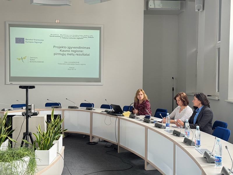 Nuotraukoje matoma konferencijų salė, kurioje trys žmonės sėdi prie apvalaus stalo, ant kurio padėti vandens buteliai, mikrofonai ir nešiojamasis kompiuteris. Priekyje rodomas projektoriaus vaizdas su informacija apie Europos Sąjungos finansuojamą projektą Kauno regione. Salėje taip pat matomi augalai ir prezentacijų įranga.