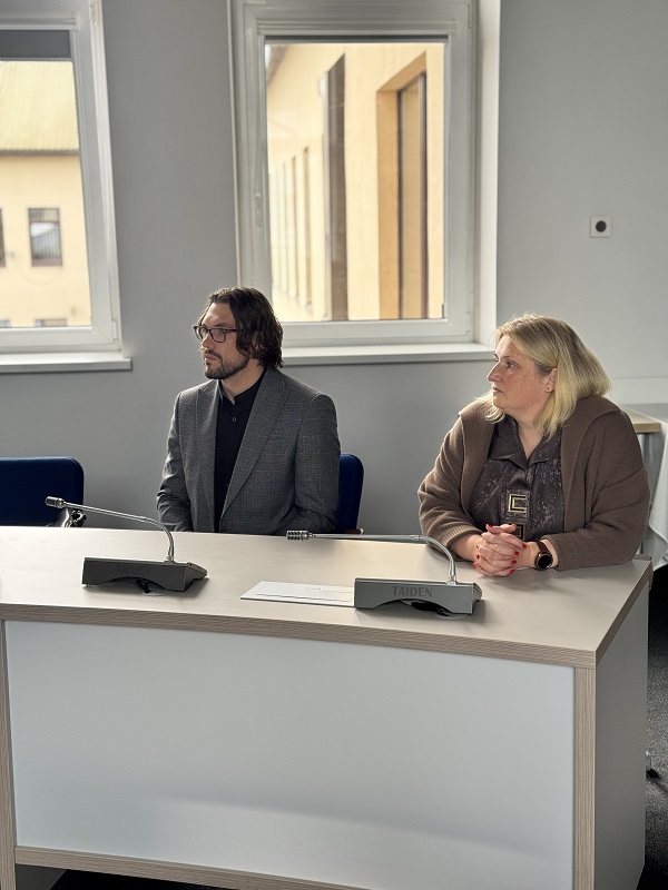 Nuotraukoje du žmonės sėdi prie stalo konferencijų ar posėdžių salėje. Ant stalo padėti du mikrofonai ir lapas popieriaus. Už jų matomi langai, pro kuriuos matosi pastatai lauke. 
