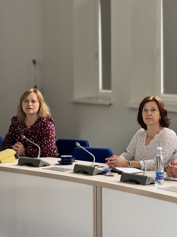 Nuotraukoje matomos dvi moterys, sėdinčios konferencijų salėje prie stalo. Prie kiekvienos jų padėti mikrofonai, ant stalo yra užrašų knygelės, mėlynas puodelis su lėkštute ir vandens butelis. Viena vilki baltus marškinėlius, kita – tamsiai raudoną palaidinę su baltais raštais. Patalpa šviesi, su langais, pro kuriuos sklinda natūrali šviesa.