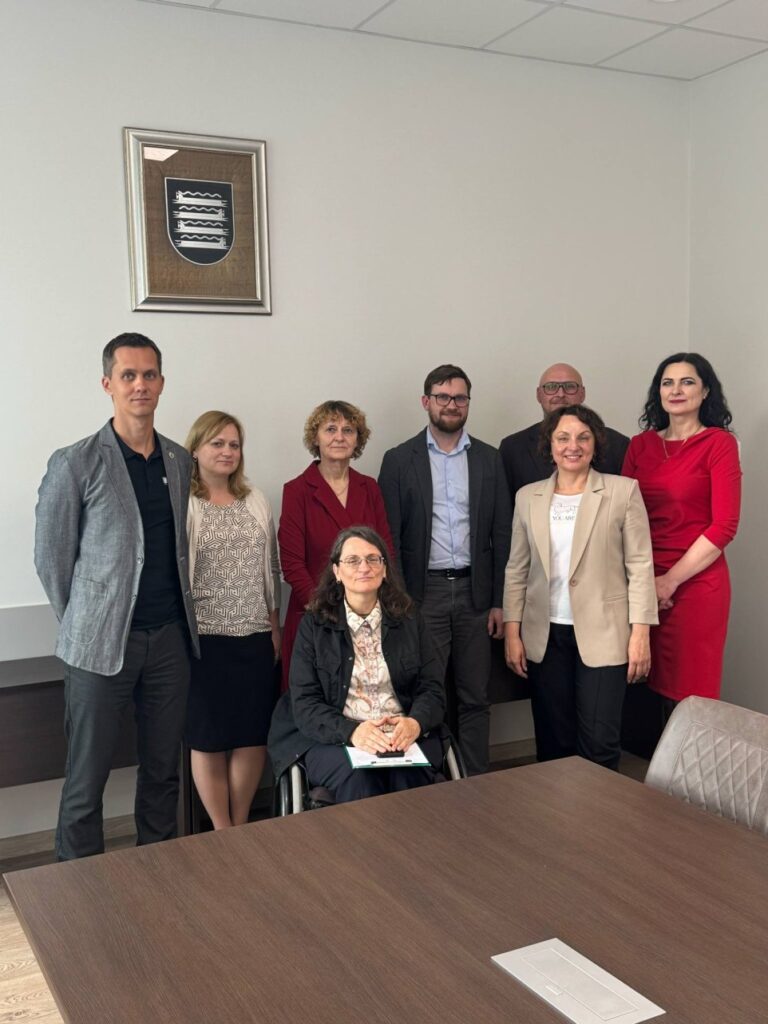 Bendra nuotrauka po susitikimo Kaišiadorių rajono savivaldybėje. Nuotraukoje matyti grupė žmonių, stovinčių patalpoje prie stalo. Patalpa atrodo kaip posėdžių ar susitikimų kambarys – ant sienos pakabintas rėmelyje herbas, o priekyje stovi didelis medinis stalas. Iš viso yra aštuoni žmonės: septyni stovi, vienas sėdi asmens su negalia vežimėlyje priekyje. Visi apsirengę tvarkingai – vyrai vilki švarkus, moterys – švarkelius, sukneles ar palaidines. Nuotaika rimta, tačiau keli žmonės šypsosi, todėl atmosfera atrodo dalykiška, bet draugiška.