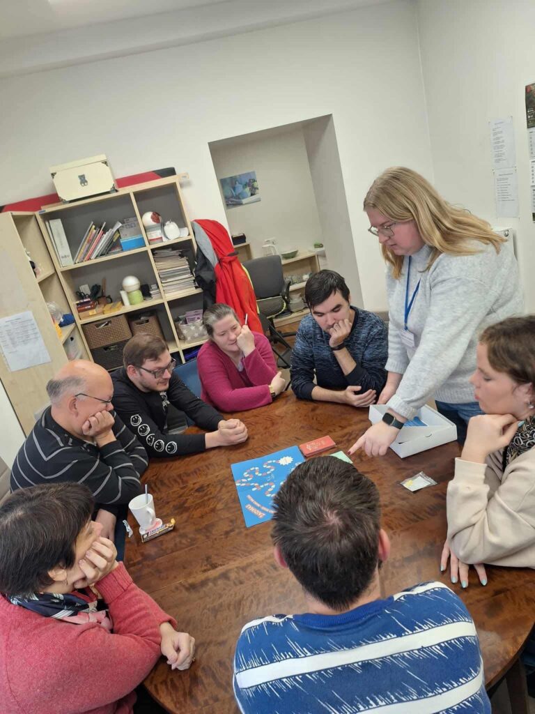 Šioje nuotraukoje matoma žmonių grupė, sėdinti aplink apvalų medinį stalą ir kartu žaidžianti stalo žaidimą.
Prie stalo aštuoni žmonės – vyrai ir moterys – o viena moteris stovi, šiek tiek pasilenkusi į priekį, ir rodo ranka į žaidimo lentą. Ji, panašu, aiškina žaidimo eigą arba padeda dalyviams atlikti ėjimą.
Ant stalo padėta mėlyna žaidimo lenta su spalvotais langeliais – matosi žodis „Žaidimas“ – bei kelios kortelių kaladės: oranžinės, žalios ir raudonos spalvos. Keli dalyviai pasilenkę į priekį ir įdėmiai stebi lentą, kai kurie – šypsosi, o kiti atrodo susikaupę.