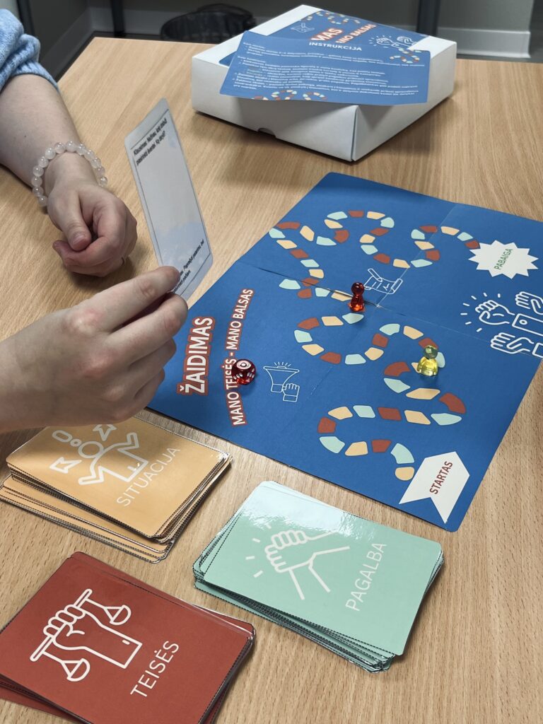 Šioje nuotraukoje matomas stalo žaidimas, padėtas ant šviesaus medinio stalo. Žaidimo lenta mėlynos spalvos, jos centre užrašyta: „ŽAIDIMAS – Mano teisės, mano balsas“. Ant lentos vingiuoja spalvotas kelias iš mažų langelių, vedantis nuo žodžio „Startas“ iki „Pabaiga“. Ant kelio stovi dvi žaidimo figūrėlės – viena raudona, kita geltona – ir raudonas kauliukas. Kairėje pusėje ant stalo išdėliotos trys kortų kaladės, kiekviena skirtingos spalvos ir temos: Oranžinė su piktograma žmogaus, rodančio į skirtingas puses – pažymėta „Situacija“. Žalsva su piktograma kumščio – pažymėta „Pagalba“. Tamsiai raudona su piktograma rankos, laikančios svarstykles – pažymėta „Teisės“. Nuotraukoje matyti žmogaus rankos. Viena iš jų laiko permatomą kortelę su tekstu, tarsi skaito užduotį ar klausimą iš žaidimo. Kita ranka ilsisi ant stalo. Žmogus, laikantis kortelę, vilki šviesiai mėlyną megztinį ir ant riešo turi šviesių karoliukų apyrankę. Fone ant stalo stovi balta žaidimo dėžė su užrašu ir sulankstytas lapas su instrukcijomis.