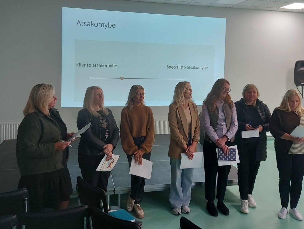 Septynios moterys stovi priešais ekraną, kuriame rodomas skaidrės užrašas „Atsakomybė“. Po juo pavaizduota linija su žymėmis „Kliento atsakomybė“ ir „Specialisto atsakomybė“.