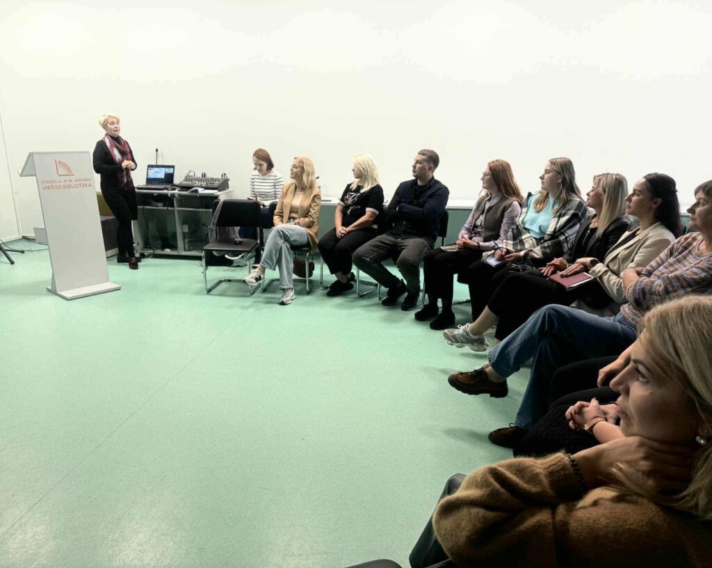 Grupė žmonių sėdi puslankiu šviesioje salėje ir klausosi kalbančios Utenos ir Panevėžio regionų tinklaveikos specialistės Tamaros Gaivoronskajos, stovinčios prie tribūnos su užrašu „Utenos A. ir M. Miškinių viešoji biblioteka“.
Fone – kompiuterinė įranga, projektorius.
