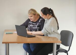 Nuotraukoje – dvi moterys, sėdinčios prie medinio stačiakampio stalo šviesiame kambaryje. Ant stalo padėtas atvertas nešiojamas kompiuteris ir maža užrašų knygutė. Abi moterys pasilenkusios prie kompiuterio ekrano: viena jų, šviesiaplaukė, vilki tamsius marškinėlius su baltu raštu, kita – tamsiaplaukė, su surištais plaukais ir pilku megztiniu – sėdi šalia. Jų laikysena rodo bendradarbiavimą ir susitelkimą – jos dirba kartu, ramiai ir įsitraukusios į užduotį. Fone – paprasta šviesi siena be papildomų detalių.