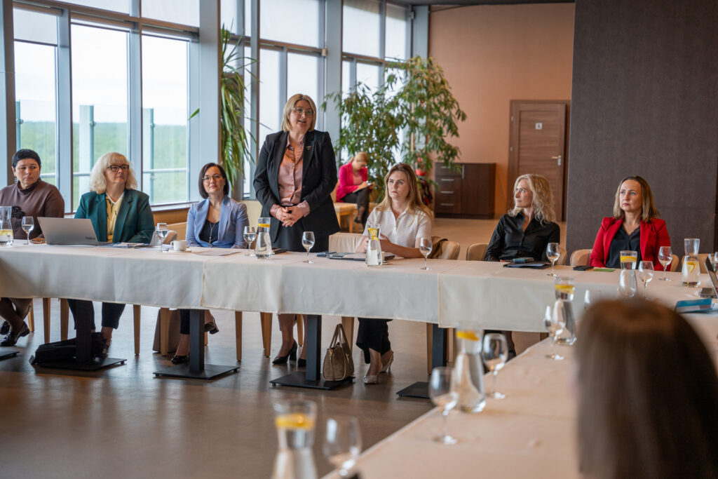 Nuotraukoje matoma grupė moterų, sėdinčių prie U formos stalo susitikimo ar diskusijos metu. Druskininkų savivaldybės mero pavaduotoją Dianą Brown, vilkinti juodą švarką ir šviesiai rausvą palaidinę, stovi ir kalba. Kitos moterys sėdi, klausosi, kai kurios turi nešiojamuosius kompiuterius ar užrašų knygas. Ant stalų stovi vandens ąsočiai, taurės ir puodeliai. Fone matosi dideli langai su vaizdu į žaliuojančią gamtą, taip pat kambaryje yra augalų, kas suteikia jaukumo.
Aplinka šiuolaikiška ir šviesi, atmosfera atrodo profesionali, bet draugiška.