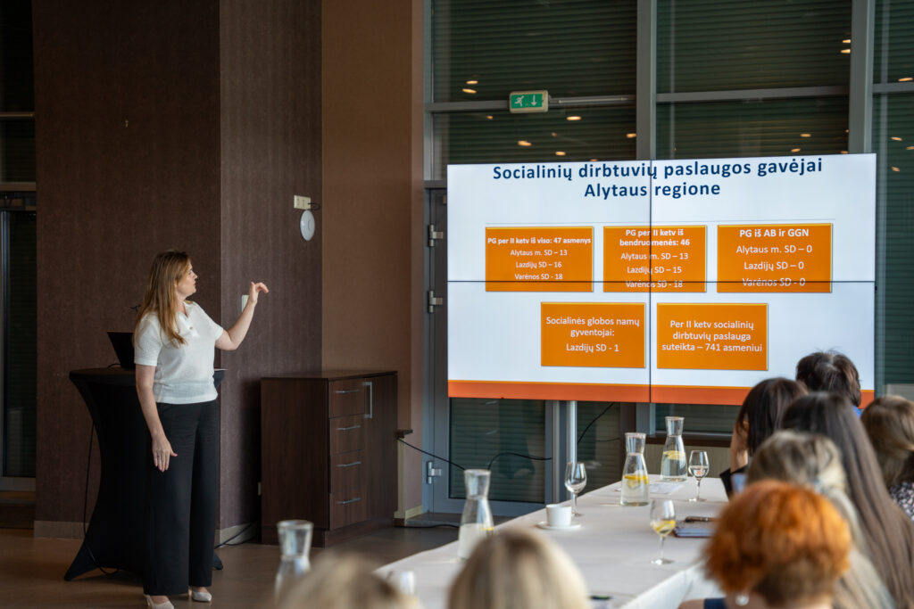 Nuotraukoje užfiksuotas pristatymo momentas. Projekto tinklaveikos specialistė Eglė Teresevičienė pristatė II ketvirčio rezultatus ir pasidalijo įžvalgomis. Eglė vilki baltą palaidinę ir juodas kelnes, stovi prie didelio ekrano, kuriame rodoma skaidrė su pavadinimu „Socialinių dirbtuvių paslaugos gavėjai Alytaus regione“. Ji rankos gestu rodo į skaidrę ir, panašu, aiškina ar komentuoja pateiktus duomenis. Ekrane matyti informacija apie paslaugų gavėjų skaičius Alytaus, Lazdijų ir Varėnos savivaldybėse. Prie stalo sėdi keli žmonės, kurie klausosi pristatymo; ant stalo stovi vandens ąsočiai, taurės, puodeliai ir užrašų knygos.
