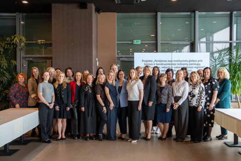 Nuotraukoje matoma grupė moterų – pozavusių bendrai nuotraukai patalpoje. Jos stovi dviem eilėmis, šypsosi ir atrodo oficialiai pasipuošusios, kai kurios vilki švarkus, sukneles ar palaidines. Aplinka – šviesi, moderni, su stiklinėmis sienomis ir žaluma fone. Dešinėje pusėje matomas stendas su tekstu lietuvių kalba, kuriame minima projekto tema:„Perėjimas nuo institucinės globos prie šeimos ir bendruomenėje teikiamų paslaugų šiaurės, vidurio ir vakarų Lietuvos regione“.